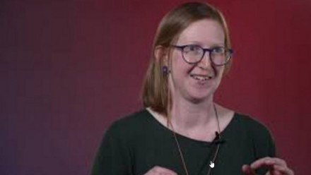A picture of Dr Bec Colvin during her TEDxCanberra speech, wearing a green dress and glasses, with a maroon background.