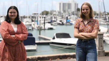 Teenagers Brooklyn O'Hearn and Claire Glavin from North Queenslad standing 1.5 metres apart, with their arms crossed, smiling and looking towards the camera. There are small ships docked at a pier behind them.