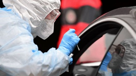 A person in full personal-protective-equipment leans into a car window to conduct a COVID-19 test swab.