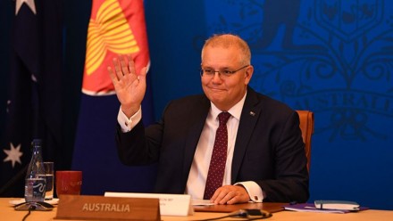 A photograph of Scott Morrison, sitting behind a desk with a plaque reading &#039;Australia&#039; at the front, raising his hand in a wave and smiling.