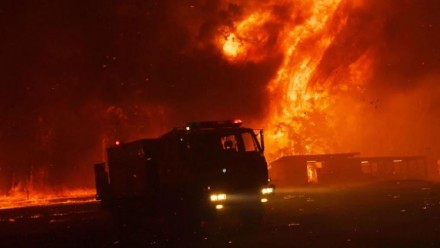 A photograph of a firetruck silhouetted against a building and surrounding landscape being engulfed in flames.