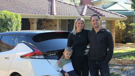 A photograph of the Dudney family standing next to their white car, in front of their beige brick house.