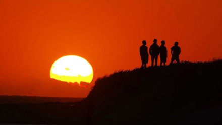 A photograph of three people standing on a hill watching the sun set.