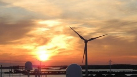 The sun sets behind a wind turbine, with a large body of water on the lefthand side.