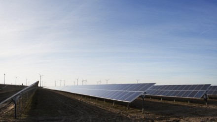 A photograph of solar panels in a field, with wind turbines in the distance.