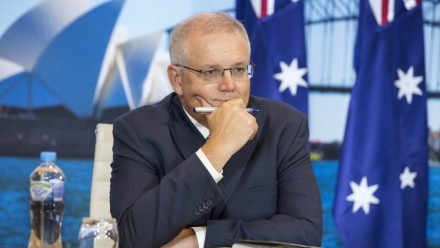 A photograph of Scott Morrison sitting at a desk looking to the right of the camera, with a picture of the Sydney Opera House and two Australian flags behind him.