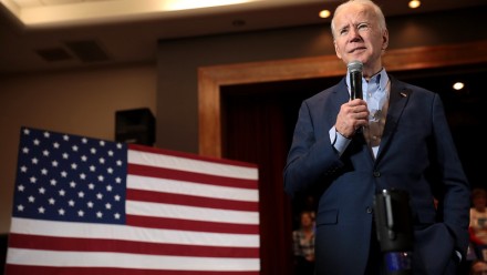 A photograph of US President Joe Biden giving a speech, with the American flag in the background.