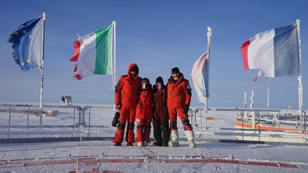 A team of four people in Antarctica, dressed in red thermal gear, standing together smiling and looking at the camera, with the French, Italian, and European Union flags behind.