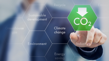 A person's hand, with a finger pointing to a green hexagon on a screen with the CO2 sign written underneath a downwards pointing arrow - indicating a decrease in carbon dioxide. Around this are other hexagons with words such as 'sustainable development'.