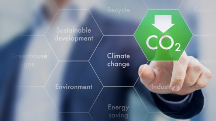 A person's hand, with a finger pointing to a green hexagon on a screen with the CO2 sign written underneath a downwards pointing arrow - indicating a decrease in carbon dioxide. Around this are other hexagons with words such as 'sustainable development'.