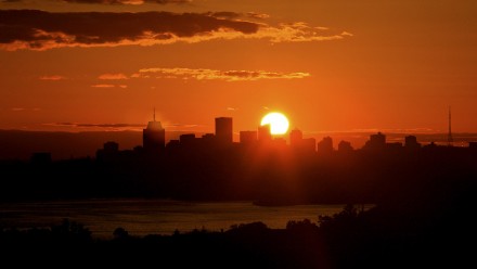 A photograph of the sun setting behind the silhouette of skyscraper towers in a city, with the sky a deep orange and hazy.