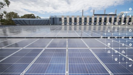 Solar panels on the roof of a building at ANU, with trees in the background and a partly cloudly sky above.