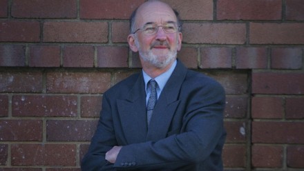 Photo of Dr Hugh Saddler in a blue suit standing with arms folded in front of a brick wall