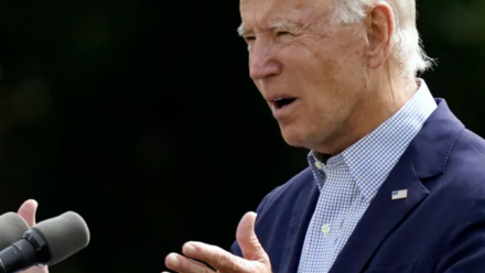 A photograph of Joe Biden delivering a speech outdoors, with both hands gesturing in front of him, and green foliage in the background.