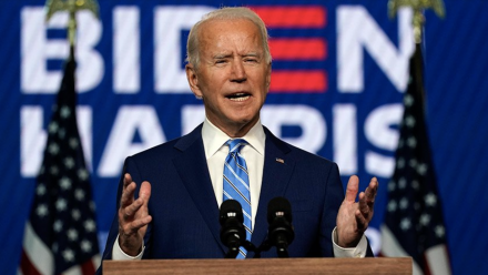 A photograph of Joe Biden delivering a speech, with the &#039;Biden - Harris&#039; presidential campaign poster in the background.