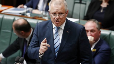 A photograph of Scott Morrison in the House of Representatives in Parliament House, standing up and talking with one finger pointing upwards.