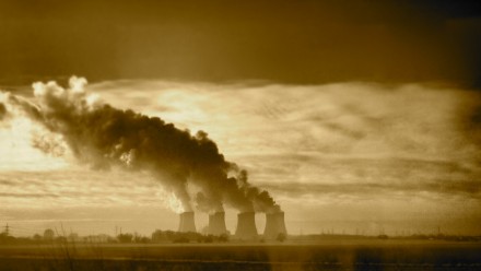 A photograph of a coal-fired power station in the distance, with the sun shining from behind, and smoke billowing out of the chimneys. 