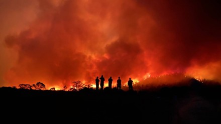 A photograph of an immense bushfire, with the silhouettes of five firefighters standing on a hill in the forefront of the blaze.