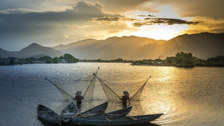 The sun sets behind mountains in the distance, with the two people fishing in the Mekong River Basin in the foreground.