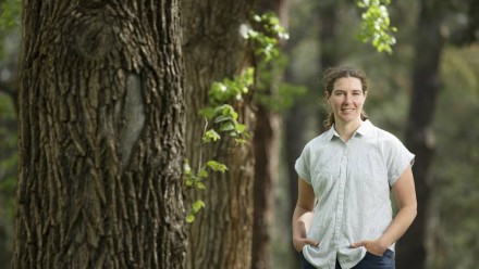 Dr Adele Morris standing next to a large tree in a forested area, looking at the camera and smiling