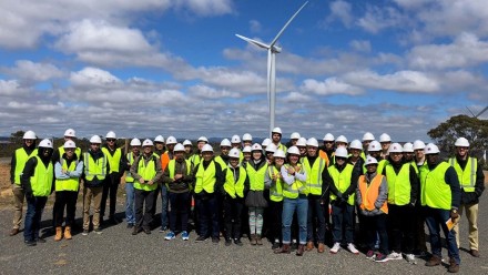 A group of students in high-vis vests and hard hats standing in front of a wind turbine.