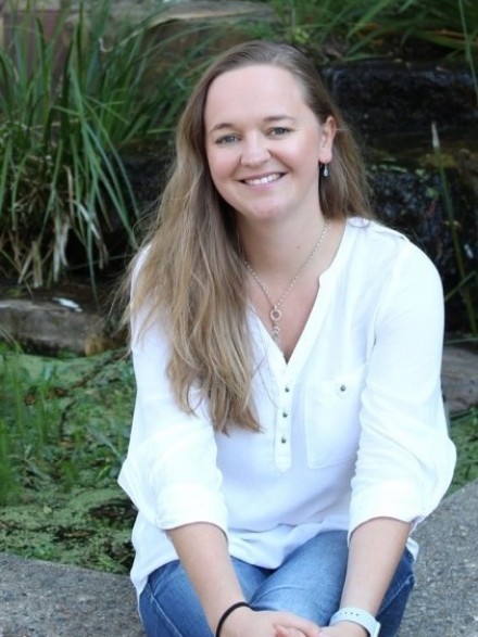 Melanie Pill seated outdoors, with green leafy plants in the background.