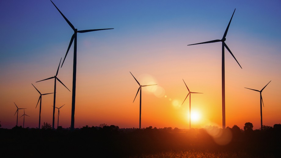 A photograph of the sun setting behind a field of wind turbines, with the sky a deep blue colour at the top, fading into orange on the horizon.