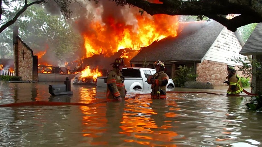 A house on fire, whilst also sitting in flood waters, with two fire fighters standing in the front.