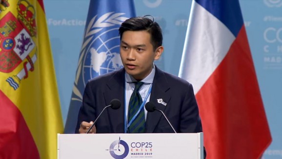 PhD student, Aaron Tang speaking at a lectern at the official Researchers and Independent NGOs statement at the COP25 with several flags in the background