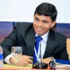 A photograph of Roshen Fernando, sitting at a desk behind a microphone, leaning forwards to speak into it.